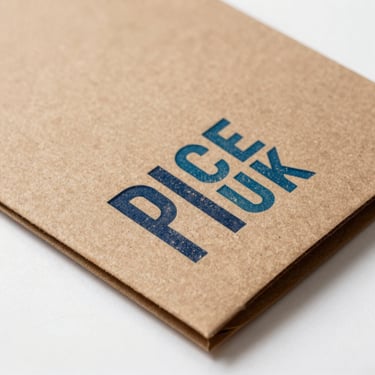 Close detail of packaging design in production — uncoated kraft board with a single-color typographic stamp, studio strobe lighting with clean shadows, shot from above on a white surface