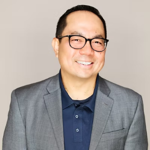 Professional corporate headshot of a smiling Asian businessman wearing glasses and a grey blazer.