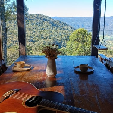 Mesa de jantar com violão em cima, xícaras de café e vista panorâmica das montanhas ao fundo em cabana Wolken Haus Urubici