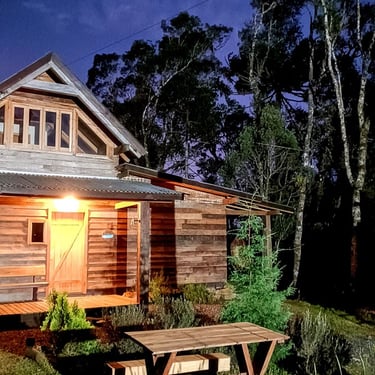 Foto noturna da frente da cabana Wolken Haus Urubici no morro da igreja com uma mesa de jardim e natureza ao redor