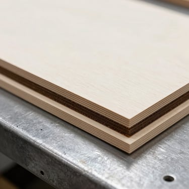 Macro shot of a laser-cut birch plywood template edge, showing clean dark-brown charred layers, resting on a steel workbench, cool workshop light.