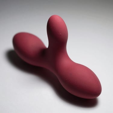 Macro studio shot of a slim G-spot vibrator with curved tip in deep red silicone on white surface, overhead light revealing smooth finish and ergonomic bend, no people