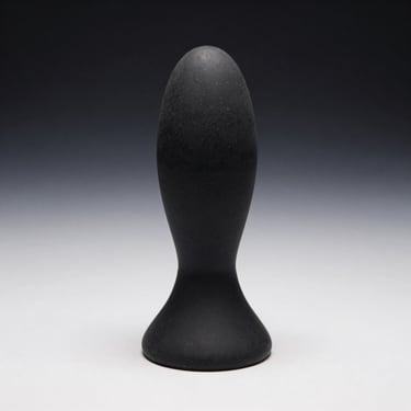 Straight-on studio shot of a vibrating anal probe with flared base in black silicone on white surface, side directional light revealing tapered shape and smooth finish, no people