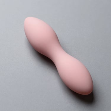 Overhead studio shot of a clitoral suction stimulator in blush silicone on grey surface, soft-angled overhead light showing nozzle detail and smooth body, no people