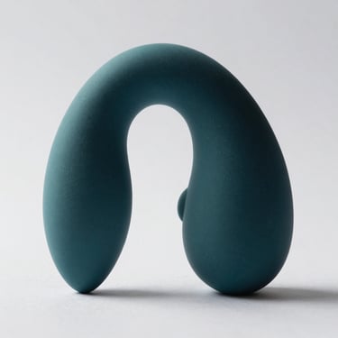 Macro studio shot of a curved prostate vibrator in dark teal silicone on white background, strong directional light from left highlighting the angled tip and material finish, no people