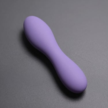Overhead studio shot of a remote-controlled wearable vibrator in soft lavender silicone on grey neutral background, directional overhead light showing wing shape and material texture, no people