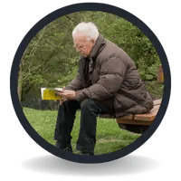 An elderly man in a brown puffer jacket sits on a park bench reading a book outdoors.