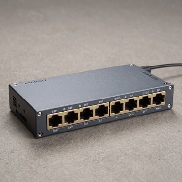 Three-quarter overhead flat-lay of a rack-mount 24-port gigabit managed switch on a neutral slate surface, studio strobe, SFP uplink ports and LED row in sharp detail, no cables