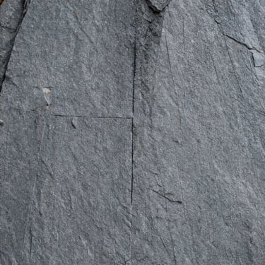 Raw quarry face of grey granite with deep vertical drill marks, cold slate tones, sharp focus.