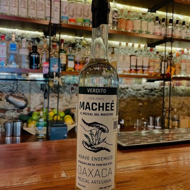 Bottle of Macheé Verdito 100% Agave Spirit sitting on a wooden bar counter, displaying the Oaxaca origin label.