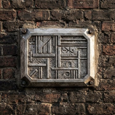 Straight-on architectural shot of an installed cast aluminum sign on a textured industrial brick wall, dramatic natural side lighting emphasizing the material weight.