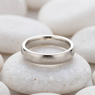 Macro shot of a sleek brushed silver band ring resting on a smooth white pebble, soft overcast daylight.