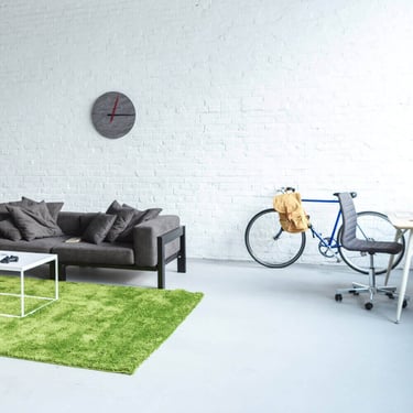 Modern studio apartment with a grey sofa, green shag rug, and blue bicycle against a white brick wall.