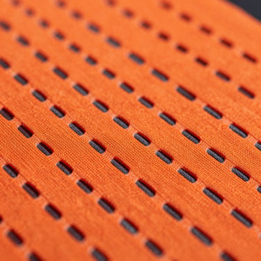 Extreme macro photography of micro-perforated athletic fabric, neon-orange and dark grey fibers woven together, dramatic side lighting.