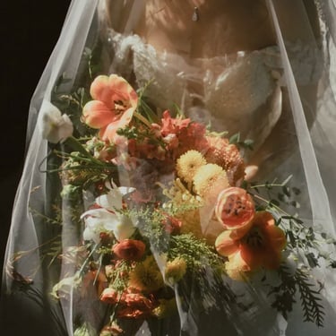 Novia con vestido de encaje bajo velo transparente sosteniendo un ramo en tonos naranja, blanco y verde.