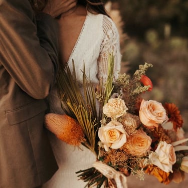 Pareja abrazada con ramo en tonos terracota y crema al atardecer.