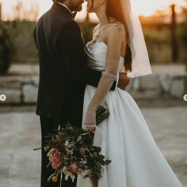 Novios abrazados al atardecer con ramo en tonos rosados.