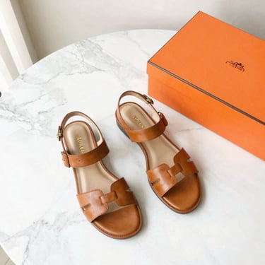 Elegant flat lay of tan leather luxury sandals next to an iconic orange box on a bright marble floor, natural daylight