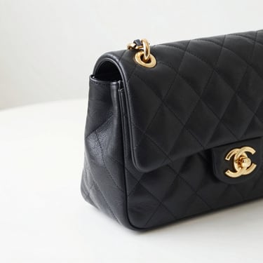 Close-up of a classic black quilted leather handbag with gold hardware, resting on a minimalist white surface, soft side lighting