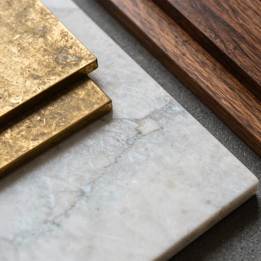 Extreme close-up detail of a raw material flat-lay, honed Arabescato marble slab next to patinated unlacquered brass and deep walnut wood samples, directional natural daylight.