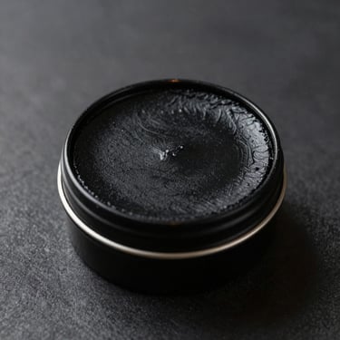 Matte black pomade tin on a brushed charcoal surface, single warm directional sidelight highlighting the lid texture, studio-controlled product shot, extreme close-up, no props