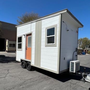 A white tiny house on wheels with a bright orange door and a mini-split AC unit parked on asphalt.
