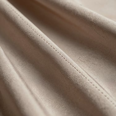 Macro detail shot of fine, precise stitching on a soft driftwood-colored silk-crepe fabric, natural daylight highlighting the texture.