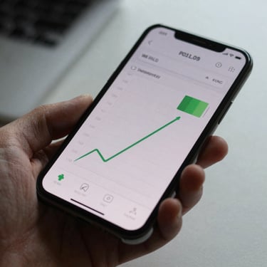 Android phone held at a natural downward angle showing a battery usage graph on screen, overcast daylight, fingers visible at the edges, plain desk surface below