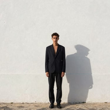 Model in full-length shot standing against a whitewashed Lisbon wall, afternoon light at 45 degrees, gaze directed just past the lens, fashion editorial framing with generous negative space above