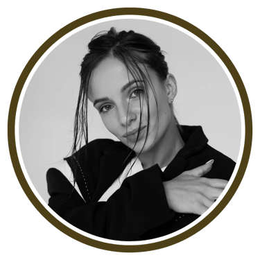 A person in a stylish black outfit poses with one arm wrapped around themselves against a muted backdrop.