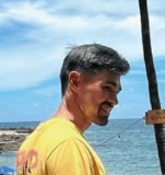 Um homem de lado, que está sorrindo, com camisa amarela, na praia.