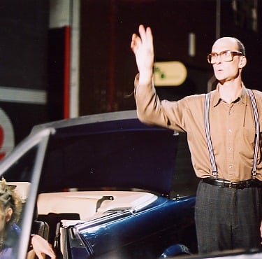 A middle-aged man wearing huge spectacles stands beside a broken-down car in which a beautiful young woman sits