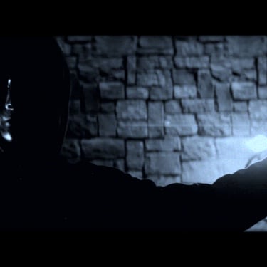 A man in a hooded sweater stands in a dimly lit room with brick walls with his outstretched arm poised to flick a wall switch
