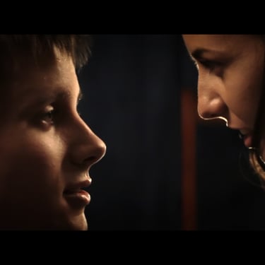 A young man/woman couple in a darkened room, their faces romantically lit by a single light that shines behind them