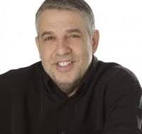 A middle aged man with greying hair, beard stubble, and wearing a brown sweater