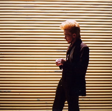 A strange little man with a 50s style hairdo stands in front of a pale-yellow corrugated roller door