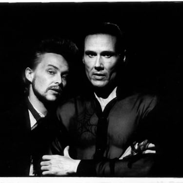 A black & white image of two men, one tall and sinister, the other shorter and sporting a beard, moustache & 50s style hairdo