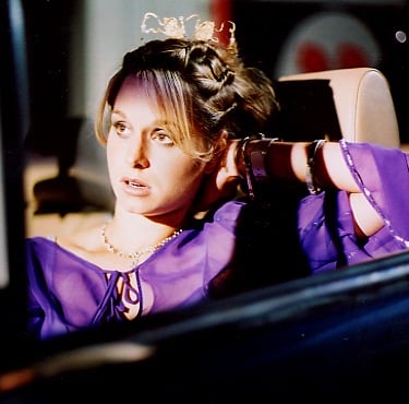 A beautiful young woman with short blonde hair and wearing a purple top, sits alone at night in a convertible car
