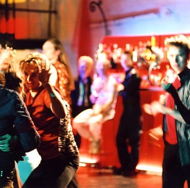 A group of young people drink at a bright red bar while other young people dance on the dance floor