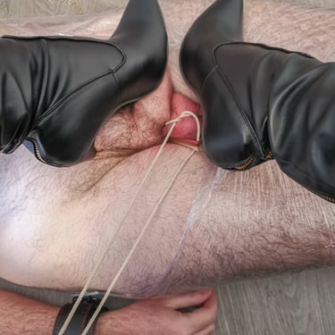 Mistress Nereid's leather-boot stilettos sink into submissive's tied testicles