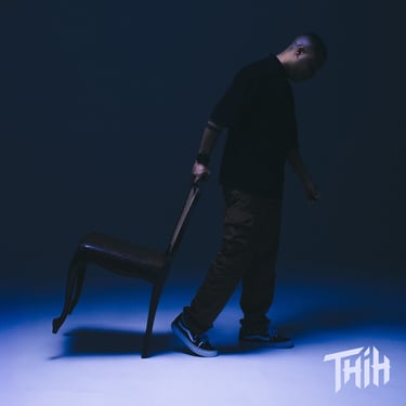 A man in streetwear pulling a chair across a blue-lit studio floor for a moody photoshoot.