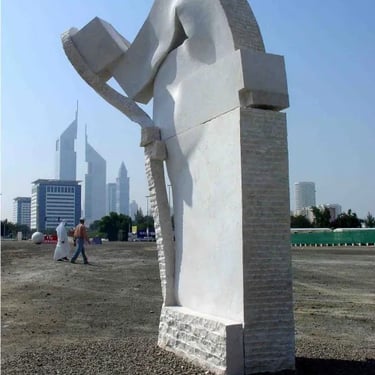 A large white stone abstract sculpture stands in a gravel lot with the Dubai skyline in the background.