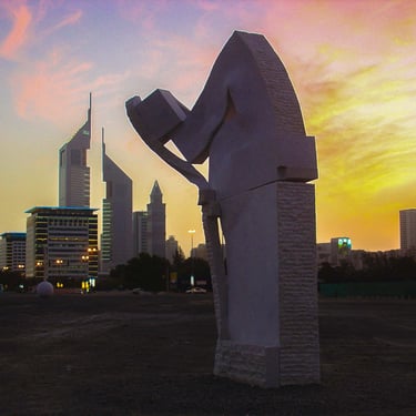 A stone sculpture stands against the Dubai skyline and Emirates Towers during a vibrant sunset.