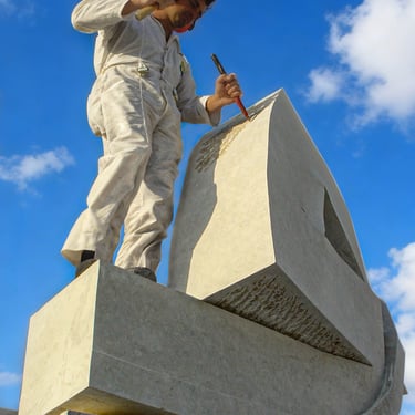 The sculptor Roberto Manzano, dressed in white, uses a hammer and chisel to sculpt a large stone monument against a blue sky.