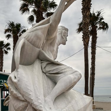 White marble sculpture of a fisherman holding a swordfish next to the beach.