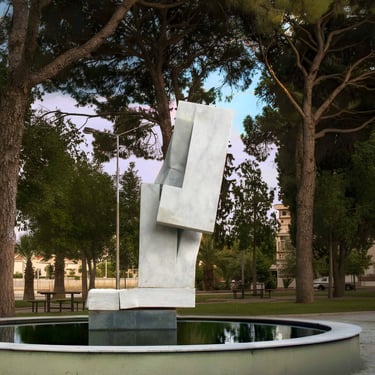 Modern white marble abstract sculpture standing in a reflective park fountain surrounded by tall pine trees.