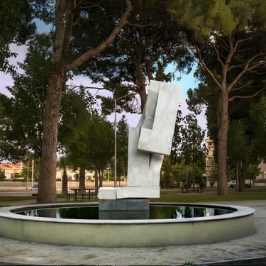 Modern white marble abstract sculpture standing in a reflective park fountain surrounded by tall pine trees.
