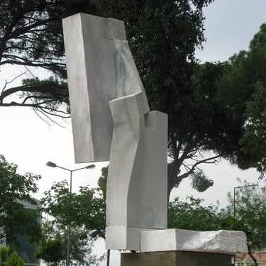 A modern abstract marble sculpture with geometric shapes standing outdoors among green trees.