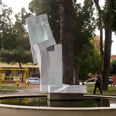 Modern white marble abstract sculpture standing in a reflective park fountain surrounded by tall pine trees.