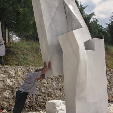 A child pushes against a large, modern white marble abstract sculpture outdoors.